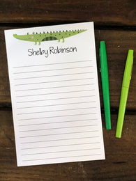 Personalized Notepad with a crocodile illustration on lined paper, personalized with a name, on a wooden surface with two green pens.